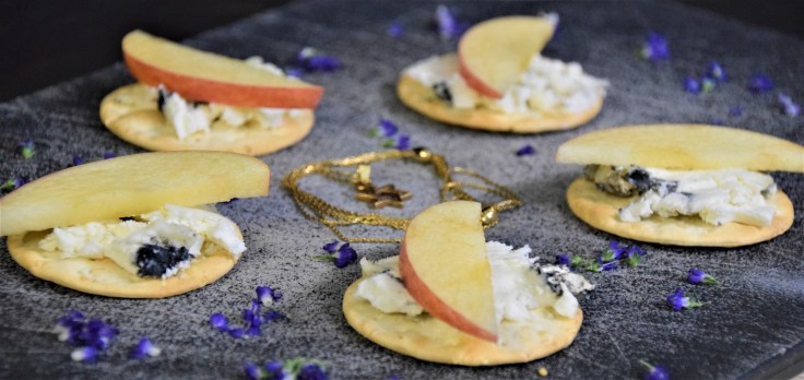 Crackers topped with white and black cheese and apple slices, circling a star necklace, surrounded by purple flower petals, all on a black background.