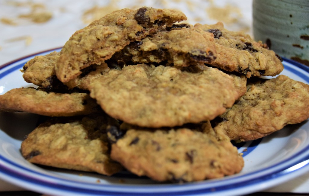 Oatmeal raisin cookies stacked on top of a white and blue striped plate; the background has a teal mug and white and gold cloth