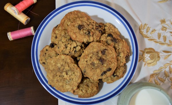 oatmeal raisin cookies on a white and blue plate surrounded by thread spools, a white and gold cloth, a wood background, and a blue mug of milk