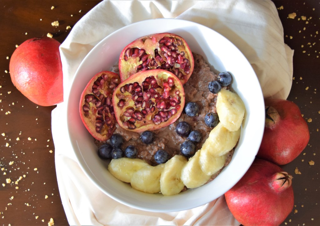 Dark pink grain porridge in a large white bowl topped with sliced pomegranates and bananas, and purple grapes. Bowl is surrounded by yellow cloth, whole pomegranates, and golden crumbs on dark wood.