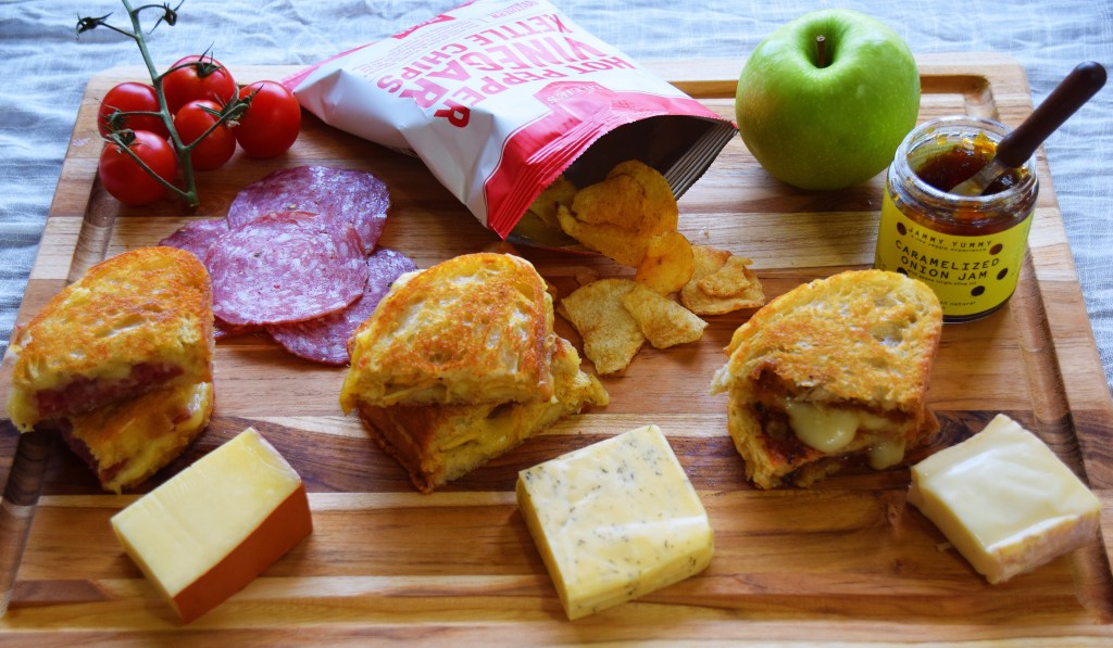 A grilled cheese themed cheese board, with three different grilled cheeses and three pale cheeses towards the bottom, a jar of onion jam and a green apple in the top right, a white and red bag of spicy chips in the top middle, and a cluster of cherry cherry tomatoes and salami in the top left.