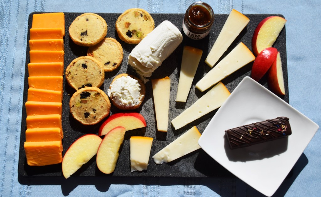 A black cheese board on a light blue background, covered by squares of orange cheddar on the left, cherry pistachio cookies, apple slices, and triangles of white gouda in the middle, a log of white goat cheese, a small jar of dark purple jam, and in the bottom right corner a white plate with a rectangular chocolate bar on it.