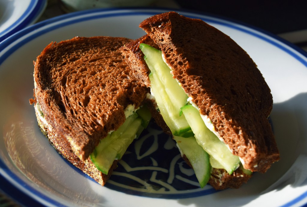 A closed face cucumber sandwich on pumpernickel bread; the sandwich is on a blue and white plate.