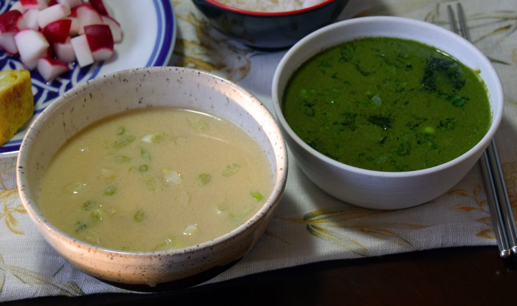 A picture of two bowls of soup, surrounded by chopsticks and plates and bowls of other food at the edges. The soup in the left bowl is pale yellow, and the soup in the right bowl is a deep green.